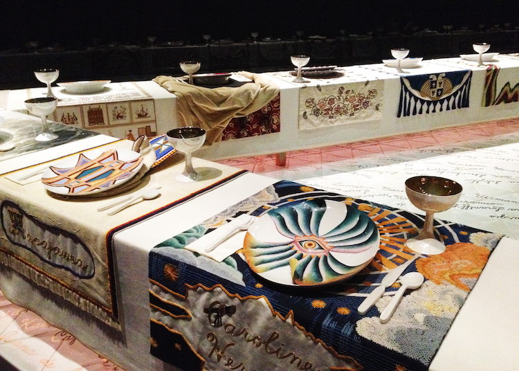 Image showing one corner of the triangular shaped table of the Dinner Party installation with 7 place settings in view each with colorful tapestry overhanging mats under ceramic plates with chalices.