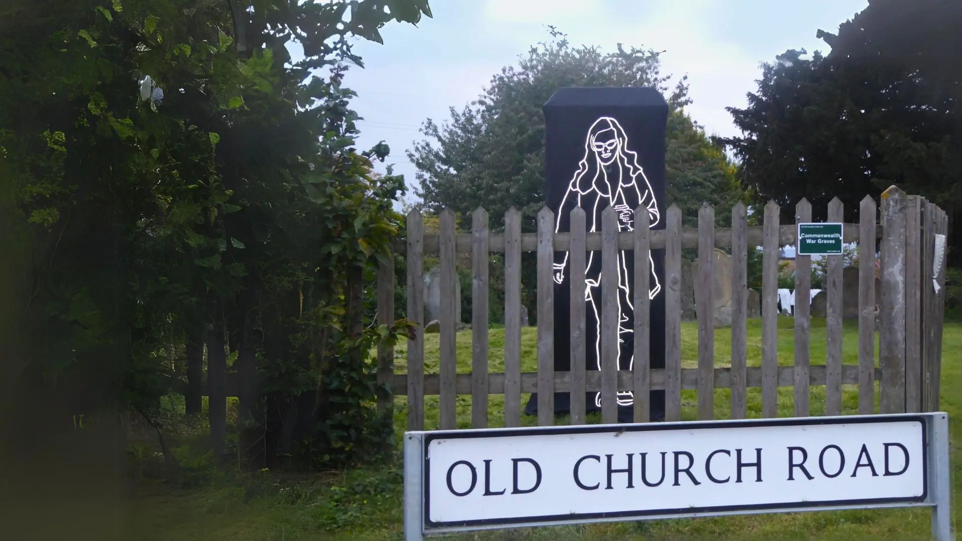 An art installation of a reflective outline of a woman walking behind a wooden fence in the grounds of a country church with a street sign in the foreground
