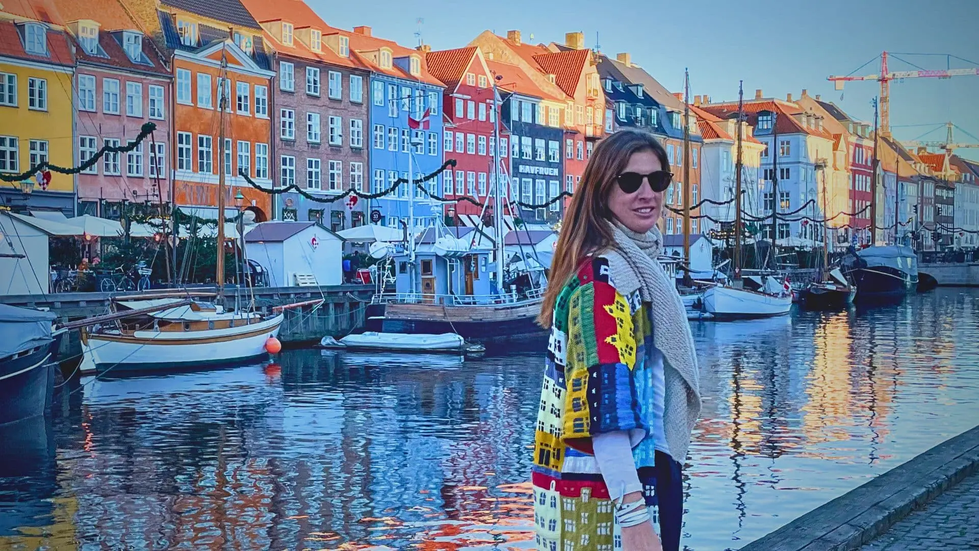 A woman standing on a dock in front of a body of water with boats and buildings in the background. She is wearing a cardigan in Jack Henzler's 'Copenhagen House Coat ' pattern. 