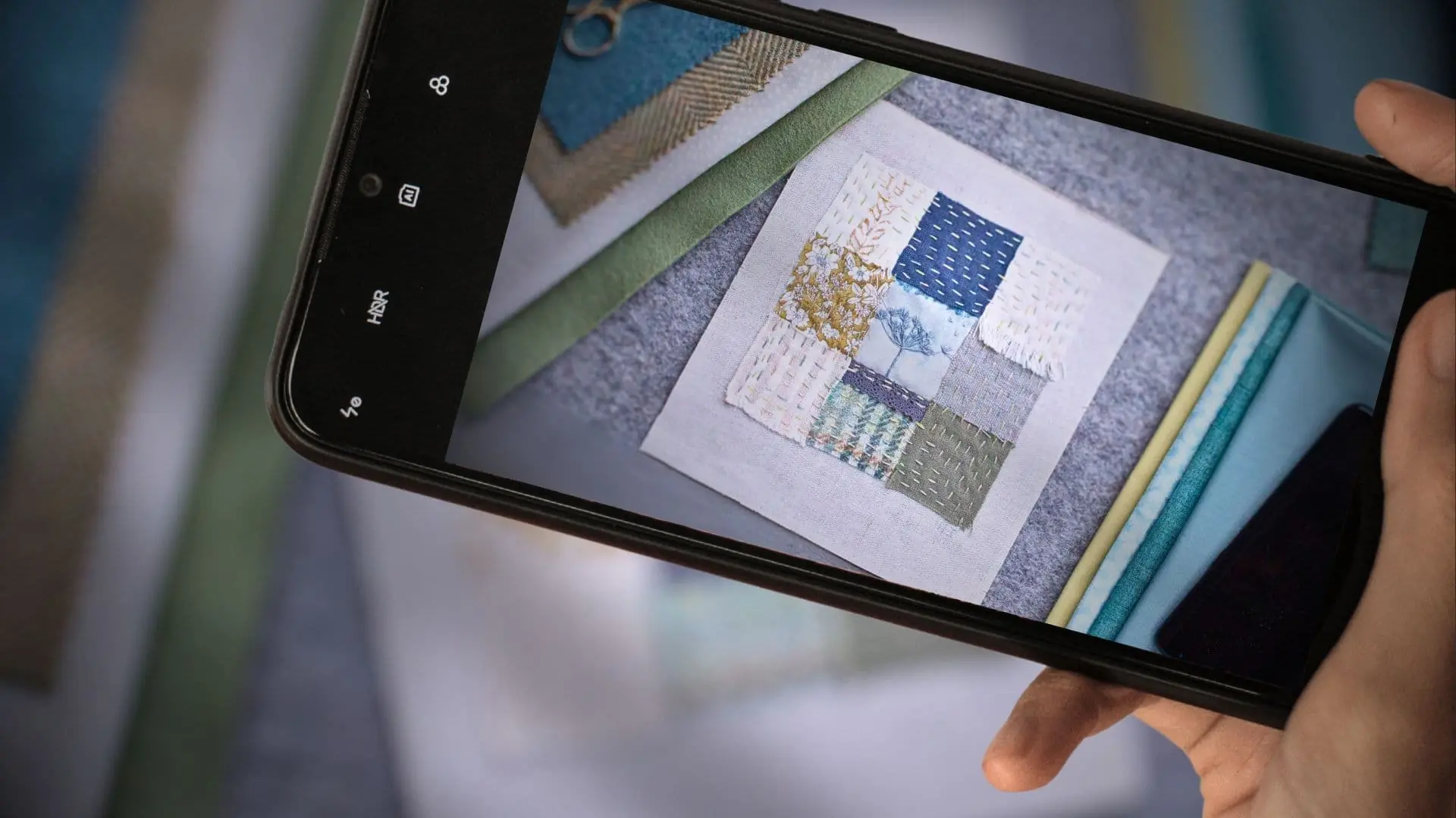 A hand holding a cell phone with a photograph of a textile art sample on the screen.