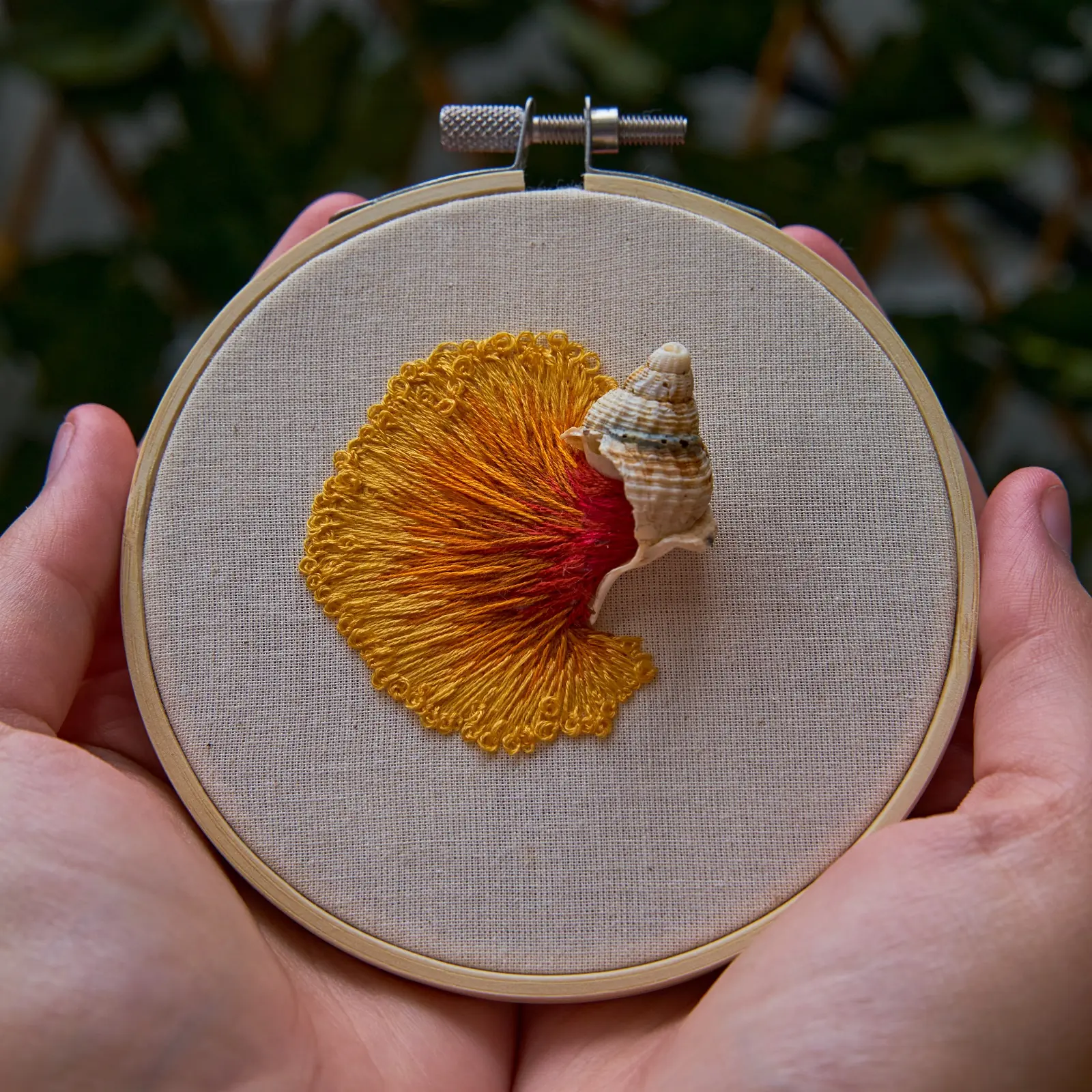 A hand holding a embroidery hoop with a seashell on it and orange thread wave coming out of it