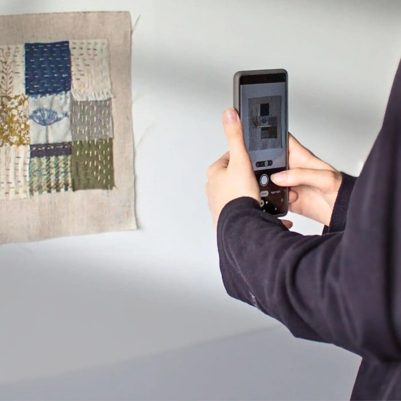 A person taking a picture of a textile art piece on a white wall