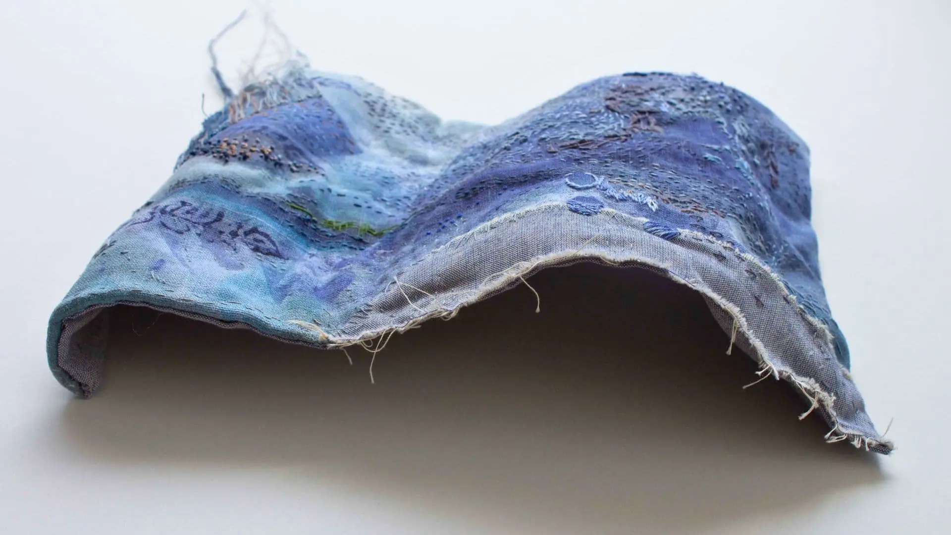 A stitched sculpture in the shape of a wave with a mix of blue colours and details and loose threads. 