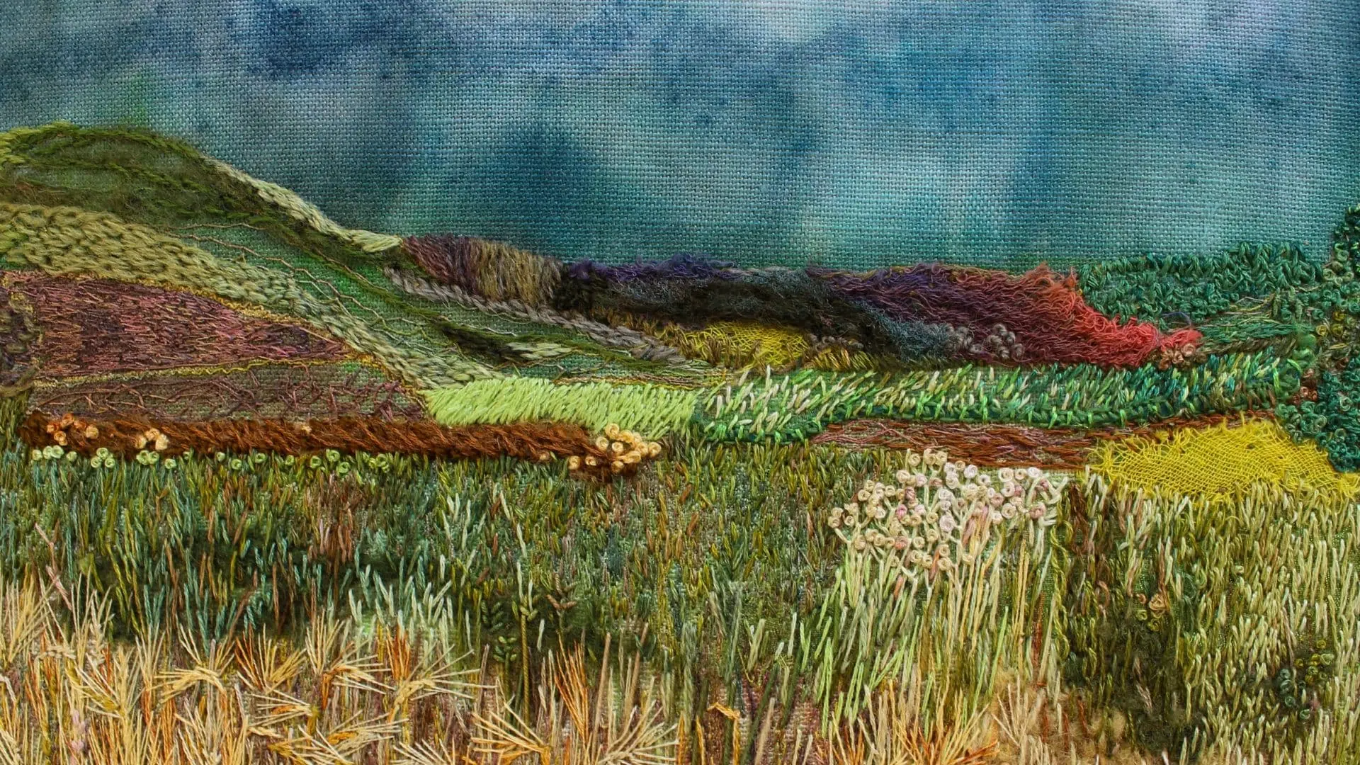 A stitched landscape with lush greens and barley fields and a brooding sky. 