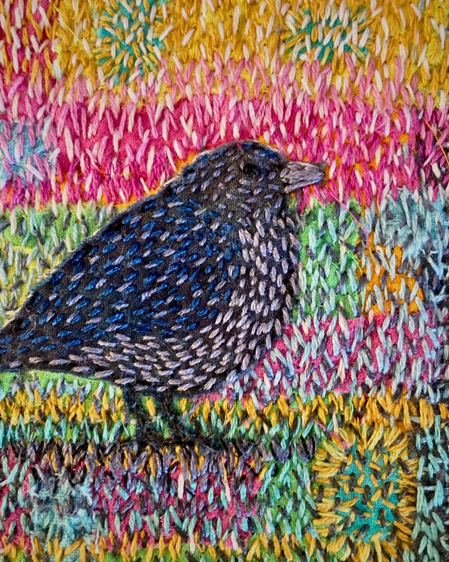Embroidered bird on colourfully stitched fabric.