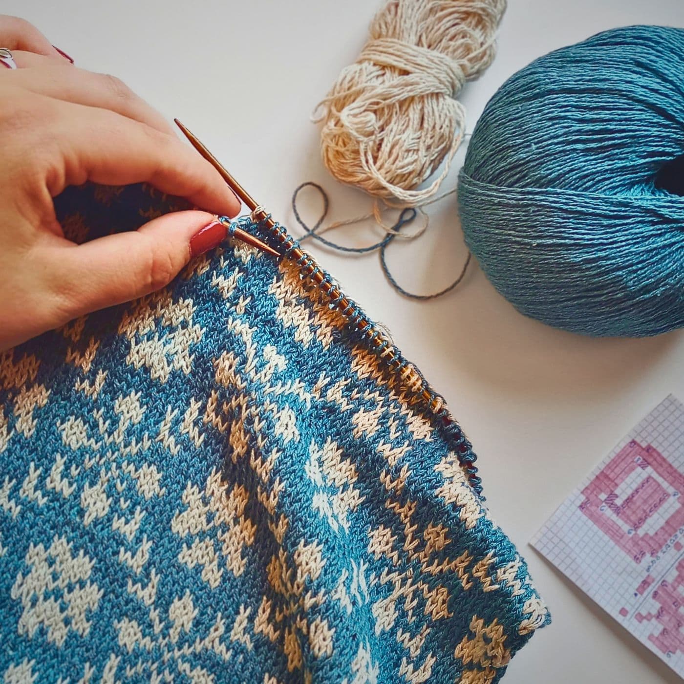 a person knitting a blue and beige pattern on a white surface.