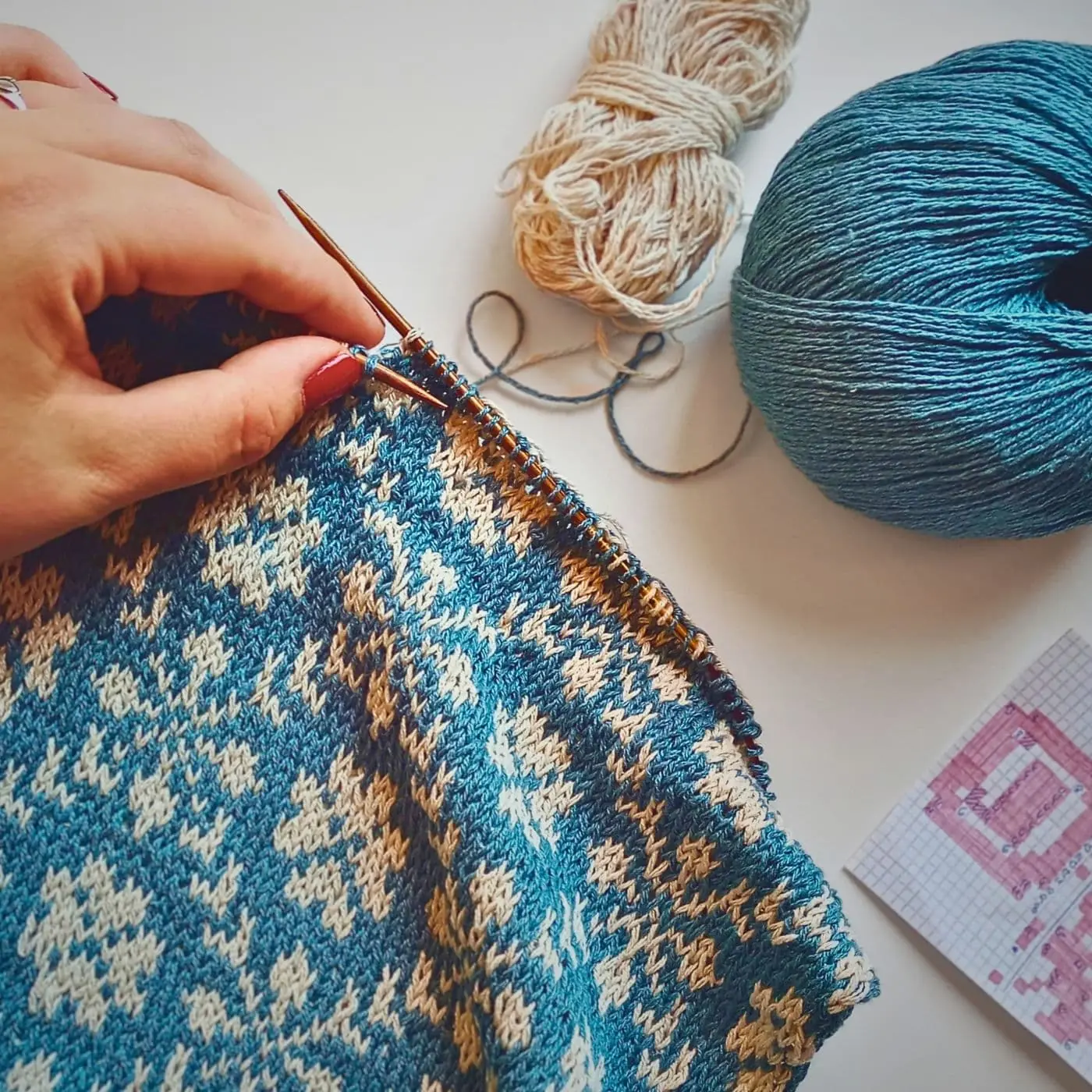 a person knitting a blue and beige pattern on a white surface.