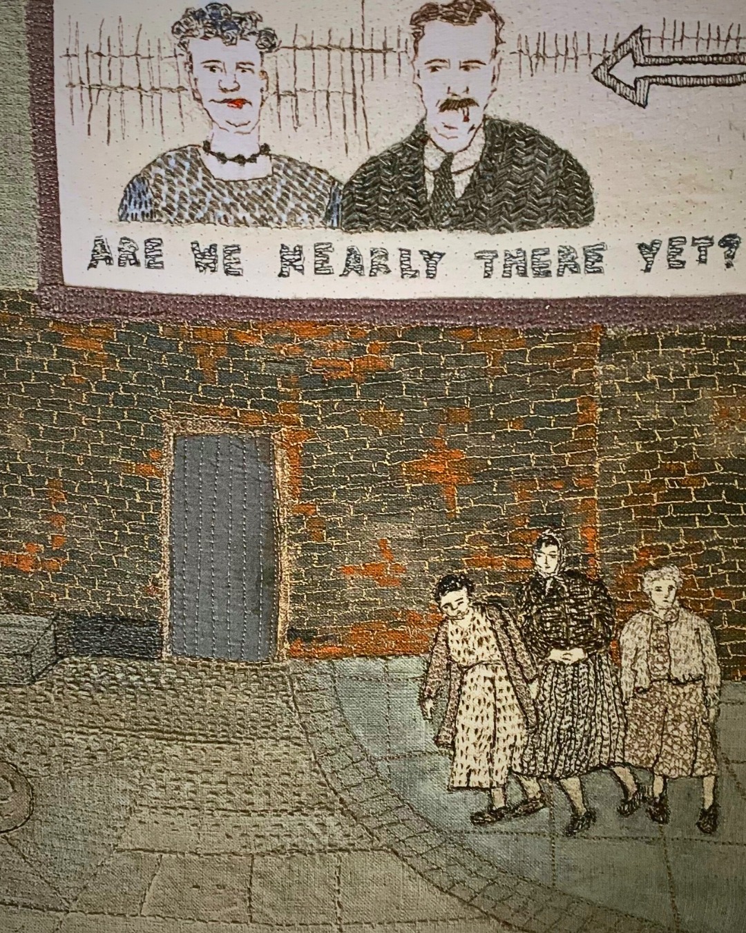 Textile art piece of women walking near a brick wall.