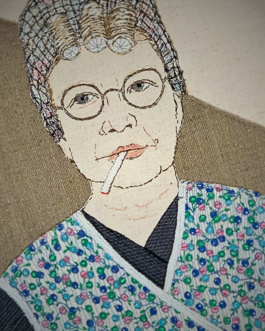 Colourful embroidered figure on fabric with hair in curlers and cigarette in mouth.